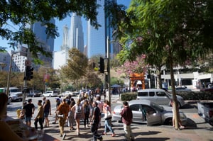 View of Downtown Los Angeles during the daytime. Tall buildings fill the scene, forming a dense city skyline. The structures vary in height and shape, with some appearing modern and made of glass, while others have more solid, rectangular designs. The perspective appears to be from street level or slightly below, looking toward the cluster of buildings. The sky is visible above, providing contrast to the vertical lines of the city.