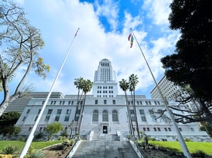 Upward view of Los Angeles City Hall from the steps of the First Street courtyard. The camera is positioned low, looking up at the tall, light-colored building as it rises into the sky. The structure has a tiered, rectangular shape that becomes narrower toward the top. The sky above is visible behind the building. The steps and lower entrance area are partially visible at the bottom of the scene, emphasizing the viewer’s position at ground level looking upward.
