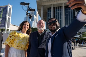 Central City Association President and CEO, Nella McOsker, poses with Homeboy Founder Greg Boyle and a guest for a selfie during the 2025 Treasures of Los Angeles Awards. They are in the forecourt of the Music Center in downtown Los Angeles. Nella is wearing a yellow and white dress, Father Boyle is wearing a clerical collar, and the guest wears a navy blue suit and sunglasses.