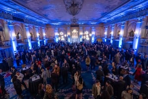 Hundreds of individuals in festive business attire gather in the ballroom of the Millenium Biltmore hotel in downtown Los Angeles for a holiday party. There are banquet tables filled with food and drinks, a stage centered in the back of the room, and holiday decor with illuminated snowflakes on the walls.