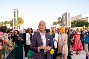 Lew Horne, wearing an eggplant-colored suit, stands in the foreground looking up toward a stage during the Central City Association’s “Treasures of Los Angeles” event. Behind him, a large crowd of attendees fills the space, many standing close together and appearing to celebrate. The scene captures a lively, high-energy moment with the audience focused on activity happening on stage.