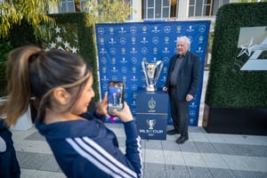A man in a suit stands in front of a step-and-repeat backdrop, posing with a trophy. He faces the camera with a composed expression while holding or standing beside the trophy. In the foreground, a woman holds up a smartphone, taking his photo.