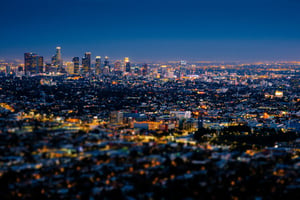 Nighttime view of downtown Los Angeles from an elevated position in Echo Park. The sky is dark blue. Below, a cluster of tall buildings fills the skyline. Many windows are lit, creating small points of yellow and orange light scattered across the buildings. The scene is viewed from above, looking down toward the city.